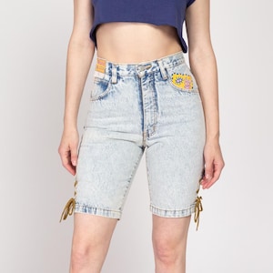 Puede incluir: Pantalones cortos de mezclilla azul claro con cordones en los lados. Los pantalones cortos tienen cintura alta y un ajuste holgado. Los pantalones cortos tienen un parche en el bolsillo trasero con un diseño colorido.