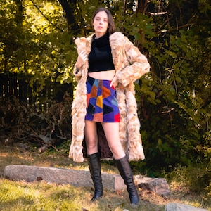 May include: A woman in a long, light brown faux fur coat, black top, and a patchwork mini skirt. She is also wearing black knee-high boots. The background is a natural outdoor setting with trees and foliage.