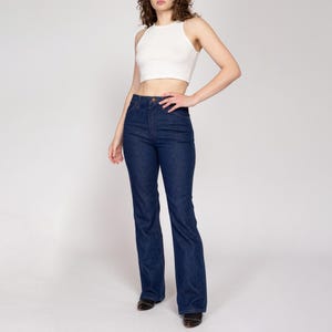 K&ouml;nnte beinhalten: Dunkelblaue Jeans mit hoher Taille und ausgestelltem Bein. Die Jeans werden mit einem wei&szlig;en &auml;rmellosen Crop-Top kombiniert. Das Model steht vor einem wei&szlig;en Hintergrund.