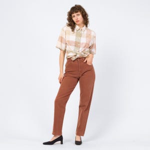 May include: A woman wearing a short-sleeved, button-down shirt with a plaid pattern in beige, pink, and green tones, tied at the waist. She is wearing high-waisted, straight-leg, rust-colored jeans and black heels.