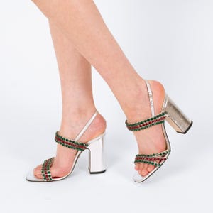 May include: Silver high-heeled sandals with a block heel. The sandals feature two straps adorned with red and green gemstones. The ankle strap is adjustable. The shoes are displayed against a white background.