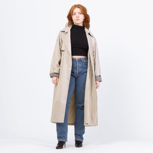 Op de afbeelding: Een beige trenchcoat met een dubbelbrede sluiting, gedragen over een zwarte coltrui en een blauwe jeans. De jas heeft een klassieke kraag, knoopsluitingen en een lange lengte. De mouwen hebben een geruite voering. De persoon draagt zwarte hakken.