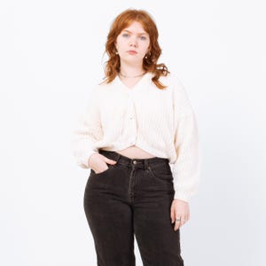 May include: Cream-colored, button-down cardigan with a textured knit pattern. The cardigan has long, voluminous sleeves and is paired with black denim jeans. The model is wearing a silver necklace and rings.