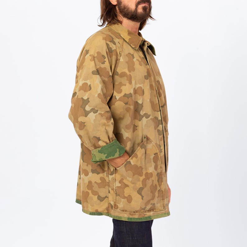 May include: A tan camouflage pattern coat with a collared neckline and two front pockets. The coat's cuffs are lined with green fabric. The coat is knee-length and has a button closure. The coat is made of a durable fabric.