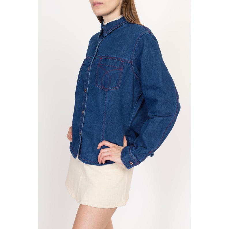 May include: A blue denim shirt with long sleeves and a button-up front. The shirt has red stitching details and a single chest pocket.