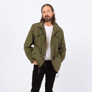 Puede incluir: Chaqueta verde oliva de estilo militar con múltiples bolsillos, cierre de cremallera y cordón en la cintura. La chaqueta se lleva sobre una camiseta blanca y combinada con pantalones negros. La chaqueta tiene un diseño clásico y utilitario.