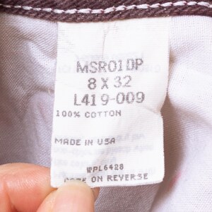 May include: Close-up of a white tag with text, including "MSR01DP", "8 X 32", and "100% COTTON". The tag is held by a person's fingers. The tag also says "MADE IN USA".