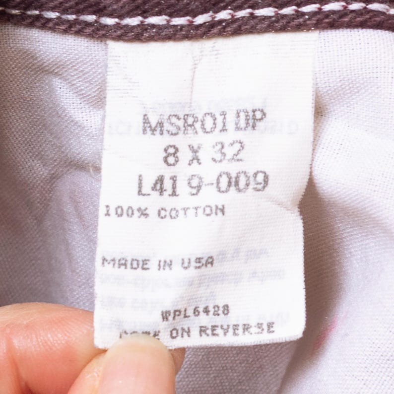 May include: Close-up of a white tag with text, including "MSR01DP", "8 X 32", and "100% COTTON". The tag is held by a person's fingers. The tag also says "MADE IN USA".