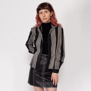 Op de afbeelding: Een zwart-wit gestreept overhemd met knopen en een zwarte turtleneck eronder. Het overhemd heeft lange mouwen en een kraag. De persoon draagt een zwarte leren rok met een ritssluiting aan de voorkant.