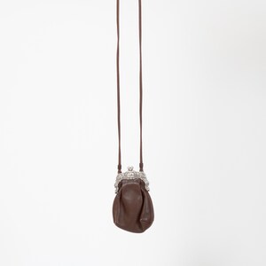 Puede incluir: Un peque&ntilde;o bolso de cuero marr&oacute;n con una correa larga y fina a juego. El bolso presenta un marco de metal plateado decorativo y ornamentado con un mecanismo de cierre de boquilla en la parte superior para el compartimento principal.
