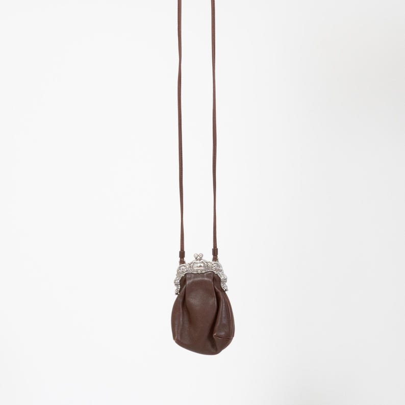 Puede incluir: Un peque&ntilde;o bolso de cuero marr&oacute;n con una correa larga y fina a juego. El bolso presenta un marco de metal plateado decorativo y ornamentado con un mecanismo de cierre de boquilla en la parte superior para el compartimento principal.