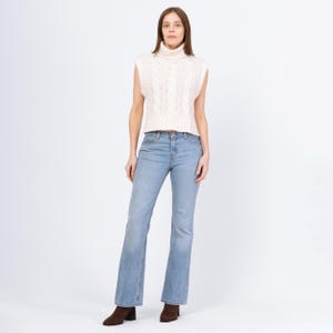 Peut inclure: Une femme porte un pull sans manches crème à col roulé en tricot torsadé et un jean bleu clair. Le jean est légèrement évasé en bas. Elle porte également des bottines marron.