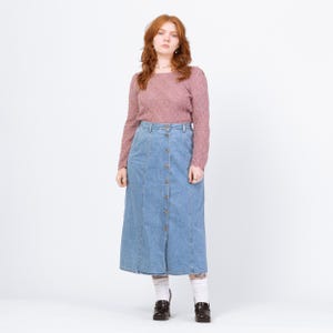 May include: A woman wearing a mauve-colored, long-sleeved sweater and a light blue denim button-down skirt. The skirt reaches the mid-calf. She is wearing black shoes and white socks with a floral pattern.