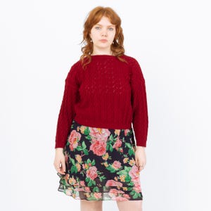 May include: A woman in a red knitted jumper and a floral skirt. The jumper has a textured pattern. The skirt features a dark background with pink and yellow roses.