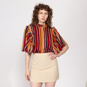 Puede incluir: Una blusa de manga corta de cuello alto a rayas rojas, naranjas, amarillas, negras y blancas con un cuello fruncido. La blusa se combina con una minifalda beige.
