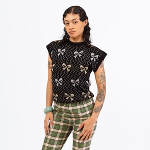 Può includere: Un maglione nero senza maniche con un motivo a fiocco dorato e argento. Il maglione è indossato sopra un paio di pantaloni a quadri verdi e neri.