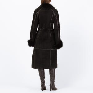 May include: A long, black shearling coat with a fur collar and cuffs. The coat features dark brown stitching and a belted waist. The model is wearing dark brown knee-high boots. A stylish winter coat.