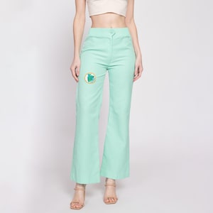 Pode incluir: Calça de perna larga verde menta com um pequeno remendo bordado na perna esquerda. O remendo apresenta um design verde, azul e amarelo.