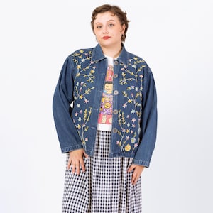 May include: A dark blue denim jacket adorned with embroidered flowers in yellow, pink, and green. The jacket is buttoned and worn over a graphic tee and a black and white checkered skirt. The jacket has long sleeves.