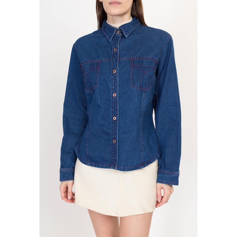 May include: A blue denim shirt with long sleeves and a button-up front. The shirt has red stitching around the pockets and collar.