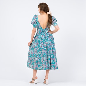May include: A teal floral print dress with a low back and short, puffed sleeves. The dress features a bow at the waist and a full skirt. The model is wearing silver heels. The dress is a vintage style.