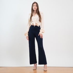 May include: A long-sleeved, light beige button-down shirt with a tied front hem paired with high-waisted, dark navy blue wide-leg trousers. The shirt features a classic collar and cuffed sleeves, while the trousers have a smooth, solid finish.