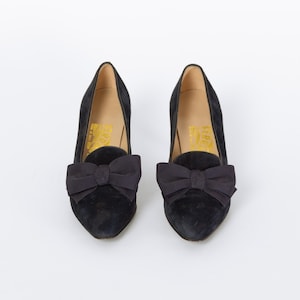 Puede incluir: Un par de zapatos de tac&oacute;n de ante negro con un lazo en la parte delantera. Los zapatos tienen un tac&oacute;n bajo y una punta puntiaguda. El interior del zapato tiene letras doradas.