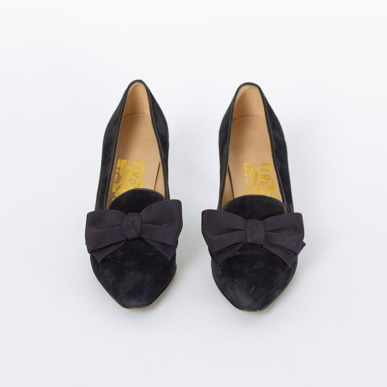 Puede incluir: Un par de zapatos de tac&oacute;n de ante negro con un lazo en la parte delantera. Los zapatos tienen un tac&oacute;n bajo y una punta puntiaguda. El interior del zapato tiene letras doradas.