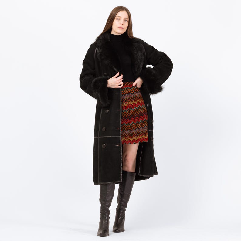 May include: A long black coat with fur trim on the collar and cuffs. The coat is buttoned and worn over a black turtleneck and a patterned mini skirt. The model is also wearing knee-high brown boots.