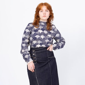 Op de afbeelding: Een blouse met lange mouwen, een hoge kraag en een donkerblauw en wit waaierpatroon. Gecombineerd met een donkere spijkerbroekrok met knoopdetails. De outfit staat tegen een witte achtergrond.