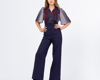 Könnte beinhalten: Eine Frau in einem marineblauen Hosenanzug. Das Oberteil ist eine gestreifte Bluse mit roten Akzenten und kurzen, ausgestellten Ärmeln. Die Plateau-Stiefel sind schwarz. Das Outfit ist vor einem weißen Hintergrund.