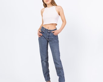 May include: A woman wearing a white crop top and blue denim jeans. She is standing with her hands in her pockets and looking at the camera.