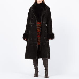 May include: A long, black coat with a double-breasted design, featuring fur trim on the collar and cuffs. The coat has dark buttons and visible stitching details. The model is wearing a patterned skirt and dark boots. The coat appears to be made of suede or a similar material.