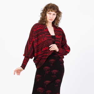 May include: A red and black patterned cardigan with long sleeves. The cardigan is paired with a black dress featuring red rose designs. The outfit is worn by a person in front of a white background.