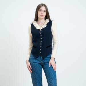 May include: A dark navy blue vest featuring a cream-colored ruffled collar, a front button closure with metallic buttons, and two horizontal slit pockets at the waist, worn over a long-sleeved cream sweater and blue denim jeans.