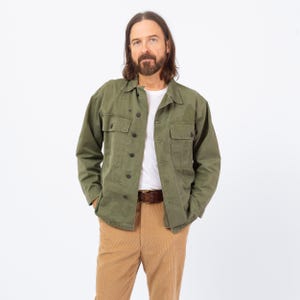 Può includere: Una giacca verde oliva con due tasche sul petto e chiusura con bottoni. La giacca è indossata sopra una t-shirt bianca e abbinata a pantaloni di velluto a coste beige e una cintura marrone. L'outfit è casual ed elegante.