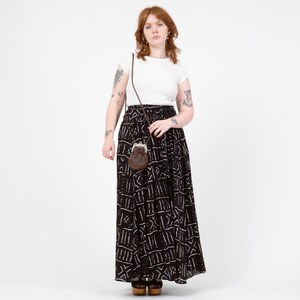 Puede incluir: Una camiseta blanca de manga corta combinada con una falda larga negra con un patr&oacute;n geom&eacute;trico blanco. La falda tiene una cintura el&aacute;stica y una silueta fluida. Un peque&ntilde;o bolso de cuero marr&oacute;n con cierre met&aacute;lico cuelga de una correa fina.