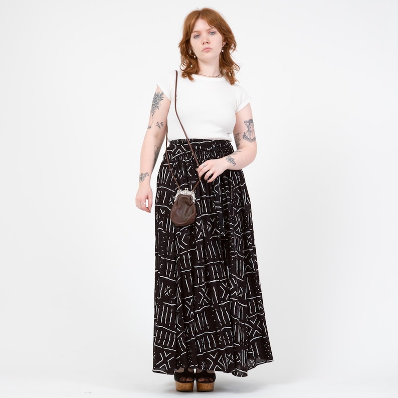Puede incluir: Una camiseta blanca de manga corta combinada con una falda larga negra con un patr&oacute;n geom&eacute;trico blanco. La falda tiene una cintura el&aacute;stica y una silueta fluida. Un peque&ntilde;o bolso de cuero marr&oacute;n con cierre met&aacute;lico cuelga de una correa fina.