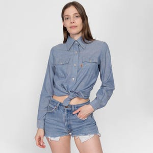 May include: A light blue denim shirt with long sleeves and a button-down collar. The shirt is tied at the waist and worn with denim shorts.