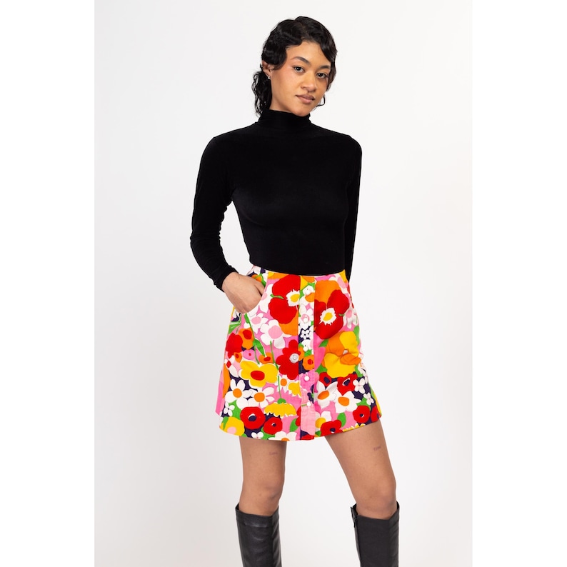 May include: A woman wearing a black turtleneck sweater and a colorful floral mini skirt with a button closure. The skirt has a pink, red, orange, yellow, and white floral pattern.