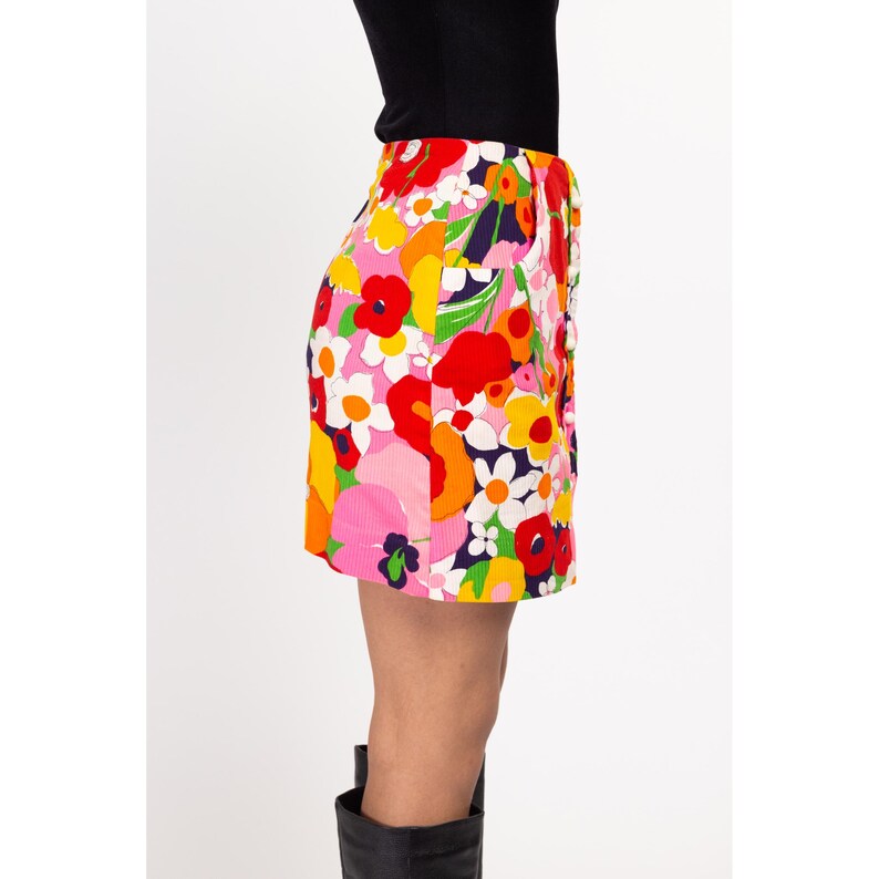 May include: A short, pink, red, orange, yellow, green, and white floral print skirt with a button closure. The skirt is made of a corduroy fabric.