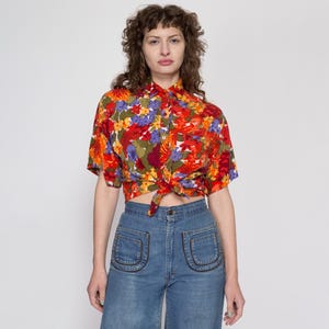 May include: A red, orange, yellow, green, blue, and purple floral print short-sleeved button-up shirt tied at the waist. The shirt is worn with blue denim jeans.