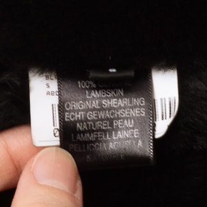 May include: Close-up of a black label with white text. The label reads "100% Genuine Lambskin, Original Shearling." The label is held against a black background, possibly a garment. The label also includes text in German and French.