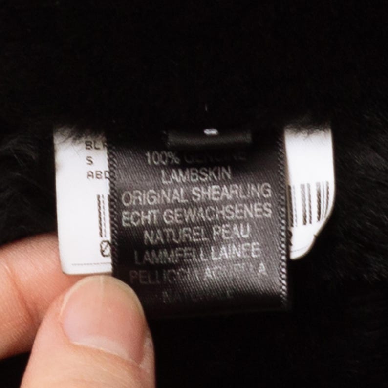 May include: Close-up of a black label with white text. The label reads "100% Genuine Lambskin, Original Shearling." The label is held against a black background, possibly a garment. The label also includes text in German and French.