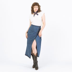 May include: A denim wrap skirt with button detailing and a high slit. The skirt is paired with a white top and cowboy boots. The outfit is set against a white background.