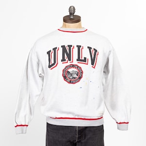 Puede incluir: Una sudadera gris con un logotipo UNLV rojo y negro y un ribete a rayas rojo y gris. El logotipo incluye las palabras "University of Nevada Las Vegas".