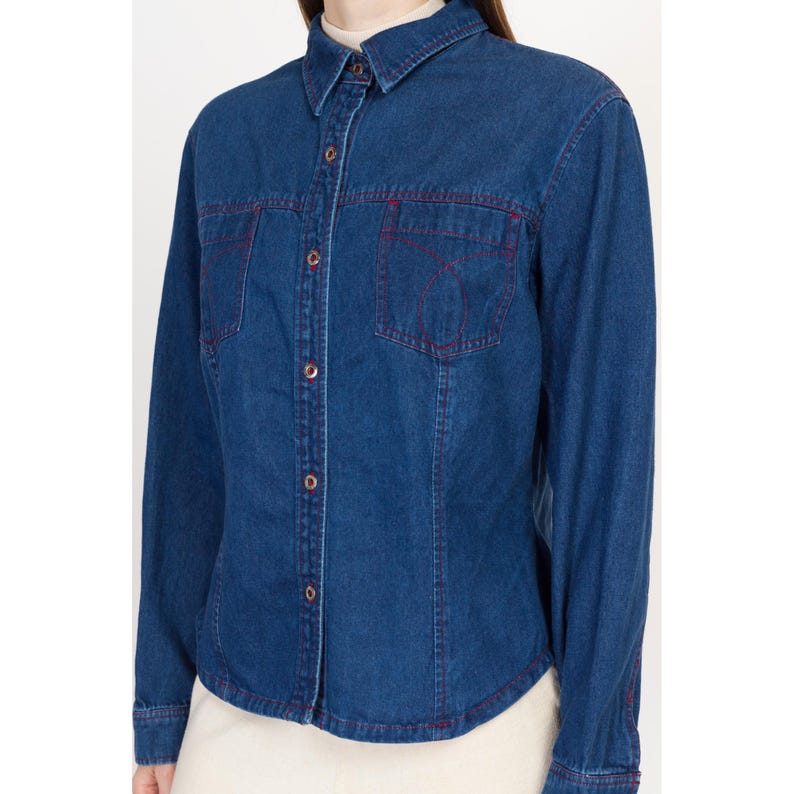 May include: A blue denim shirt with long sleeves and a button-up front. The shirt has red stitching around the pockets and collar.