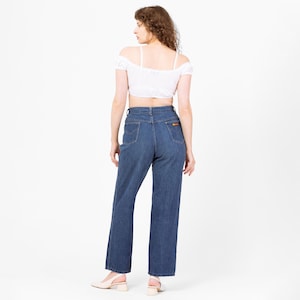 May include: A person wearing a white off-the-shoulder crop top and high-waisted, dark wash blue jeans. The jeans have a small orange patch on the back pocket. The person is wearing light-colored heels.