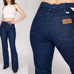 K&ouml;nnte beinhalten: Dunkelblaue Wrangler Jeans aus Denim. Die Jeans haben eine hohe Taille und ein ausgestelltes Bein. Die Ges&auml;&szlig;tasche hat einen braunen Lederaufn&auml;her mit dem Wrangler-Logo. Die Jeans sind ein klassischer Stil.