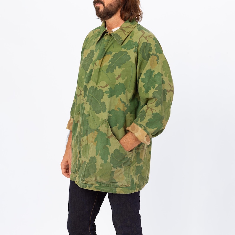 May include: A green camouflage jacket with a leaf pattern. The jacket has a collared neckline, long sleeves, and two front pockets. The cuffs are a contrasting tan color. The person is wearing dark blue jeans.